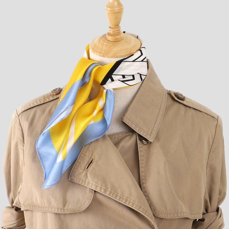 Yellow and blue geometric silk scarf knotted at the neck of a dress form wearing a beige trench coat
