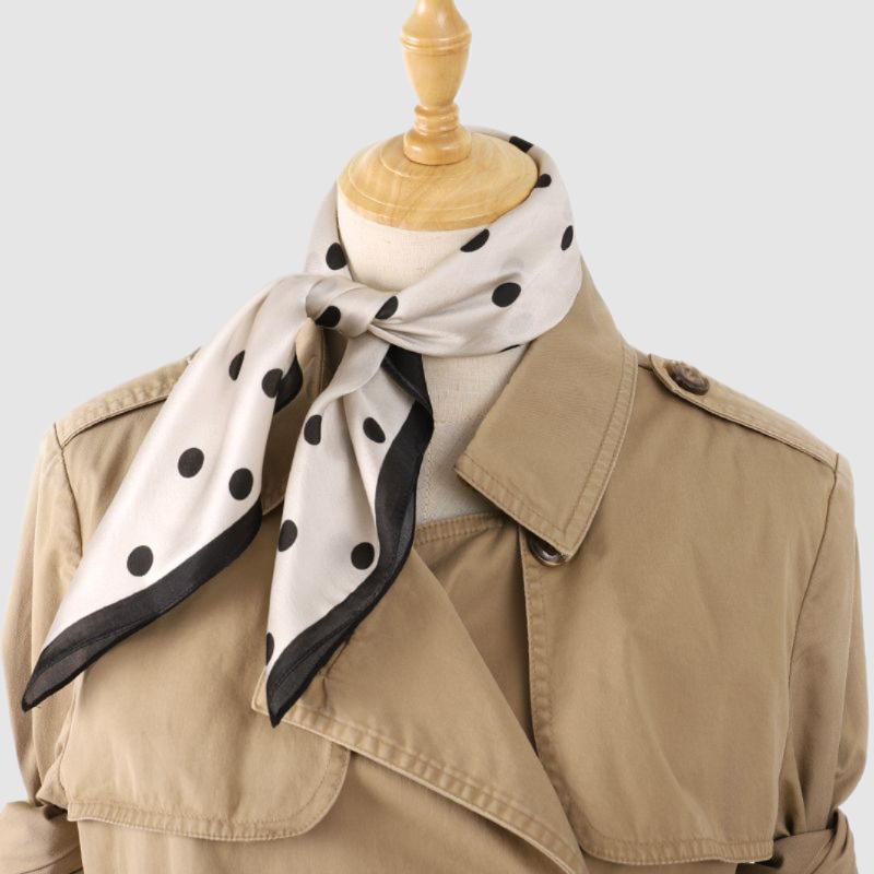 White and black polka dot silk scarf with black border, knotted at the neck of a mannequin in a trench coat