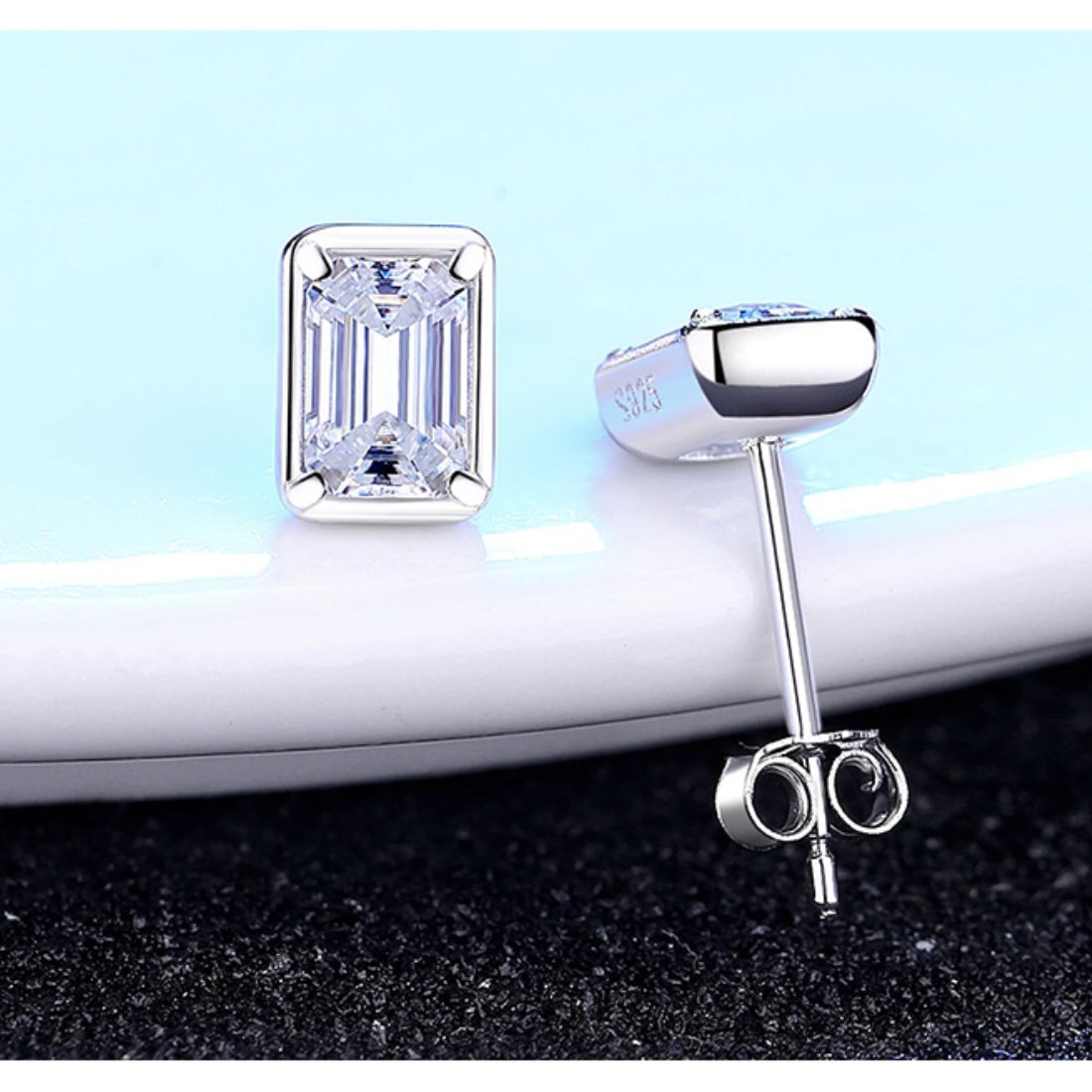 Rectangular stud earrings propped on a pale blue dish above a black glitter surface, showing front and post